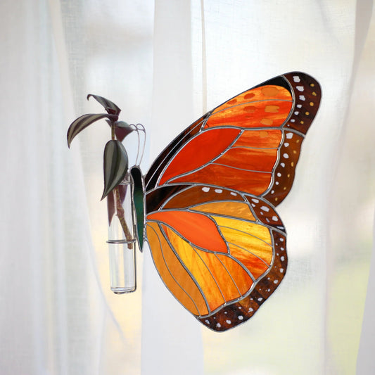 A stained glass monarch butterfly ornament with orange, yellow, and brown wings, attached to a small clear glass vase with dark green leaves, set against a white curtain background.