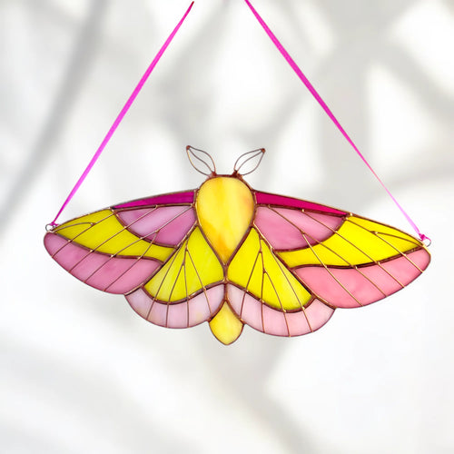 A stained-glass window hanging moth ornament with pink and yellow wings, suspended by a bright pink string against a softly lit white background.