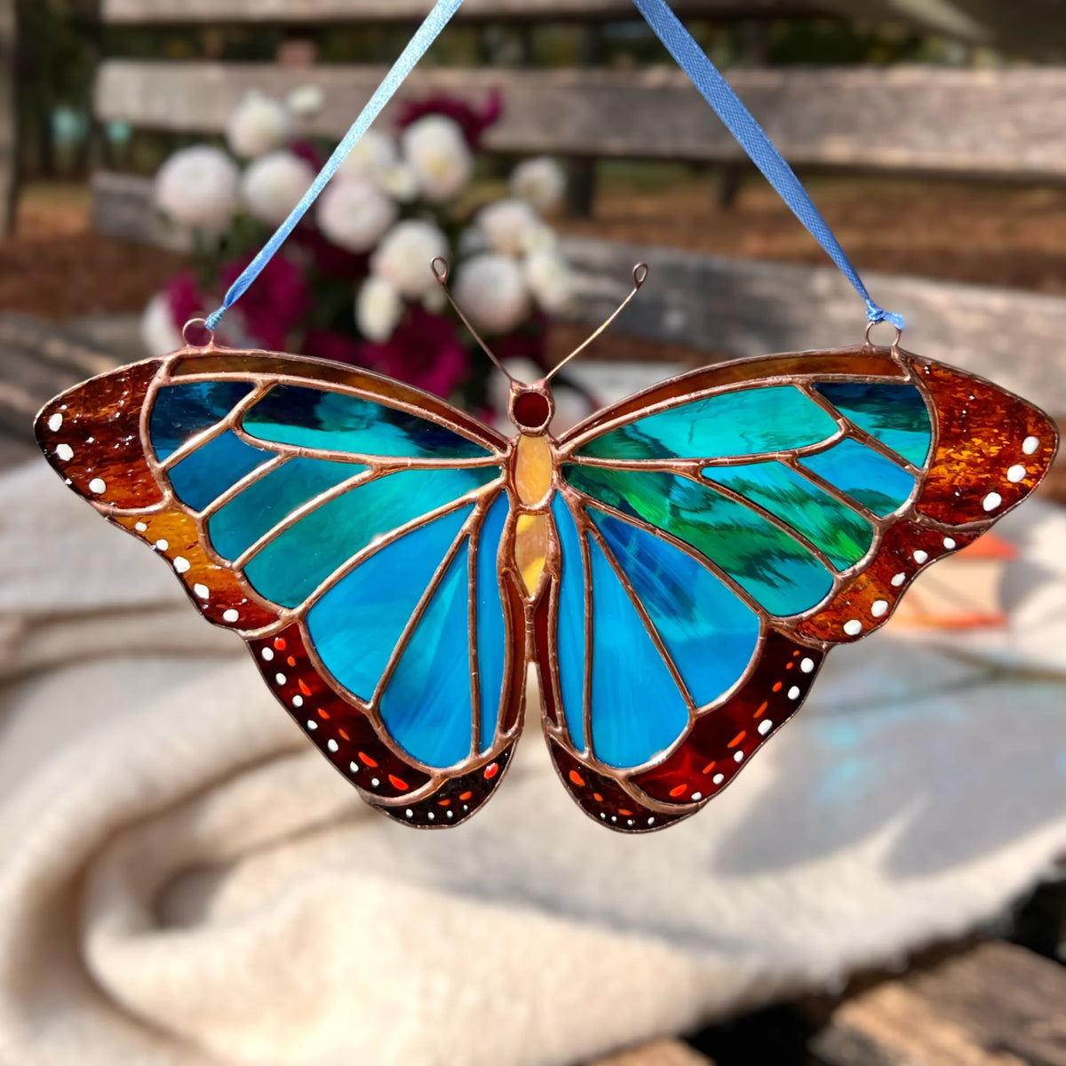 A stained glass butterfly ornament with blue stained glass wings and a blue hanging ribbon, photographed outdoors against a softly blurred garden background.