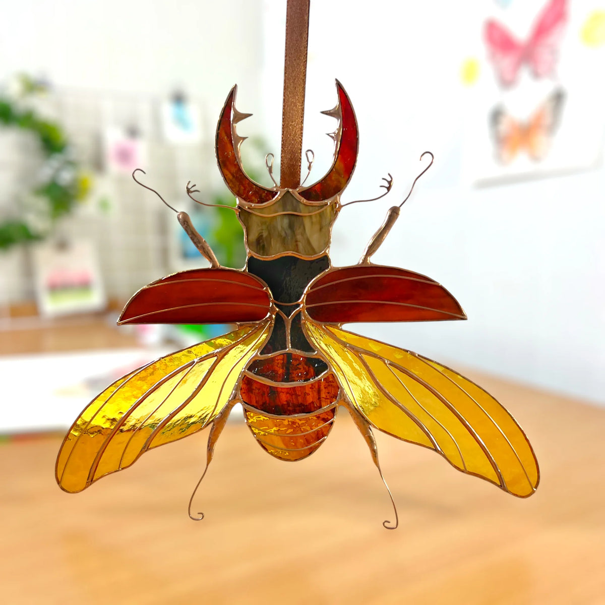 A stained glass beetle ornament hanging from a wooden dowel, featuring amber yellow wings, reddish-brown horns, and a gold-toned body, set against a softly blurred indoor background.