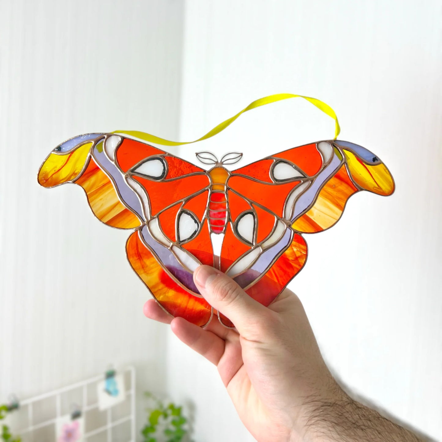 A hand holds a stained glass moth suncatcher with orange, yellow, red, and purple panels and a yellow hanging ribbon, set against a softly blurred indoor background.