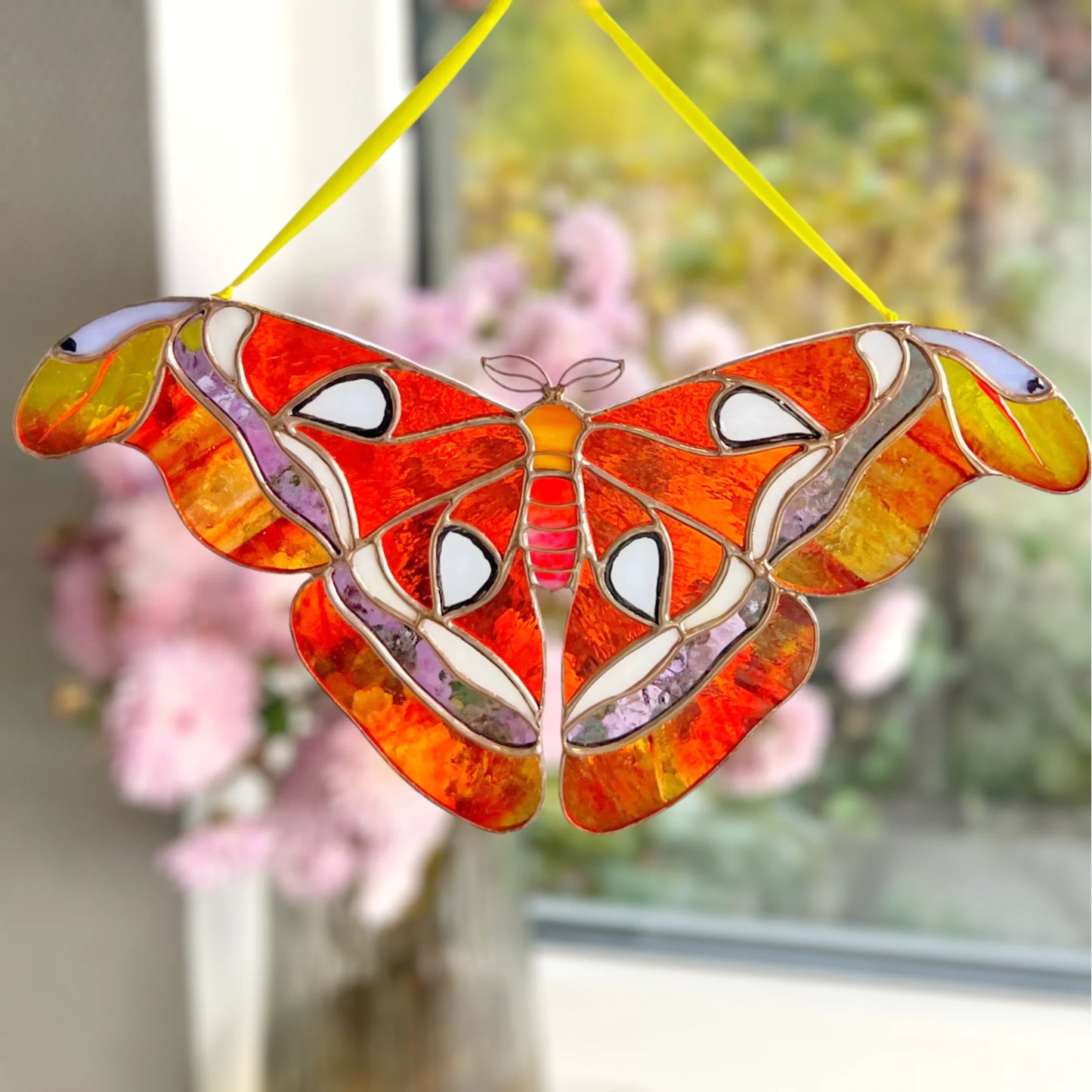A stained glass suncatcher shaped like a giant moth or butterfly, featuring vibrant orange, red, yellow, and white patterns with a yellow hanging string, shown hanging in front of a softly blurred window with pink flowers.