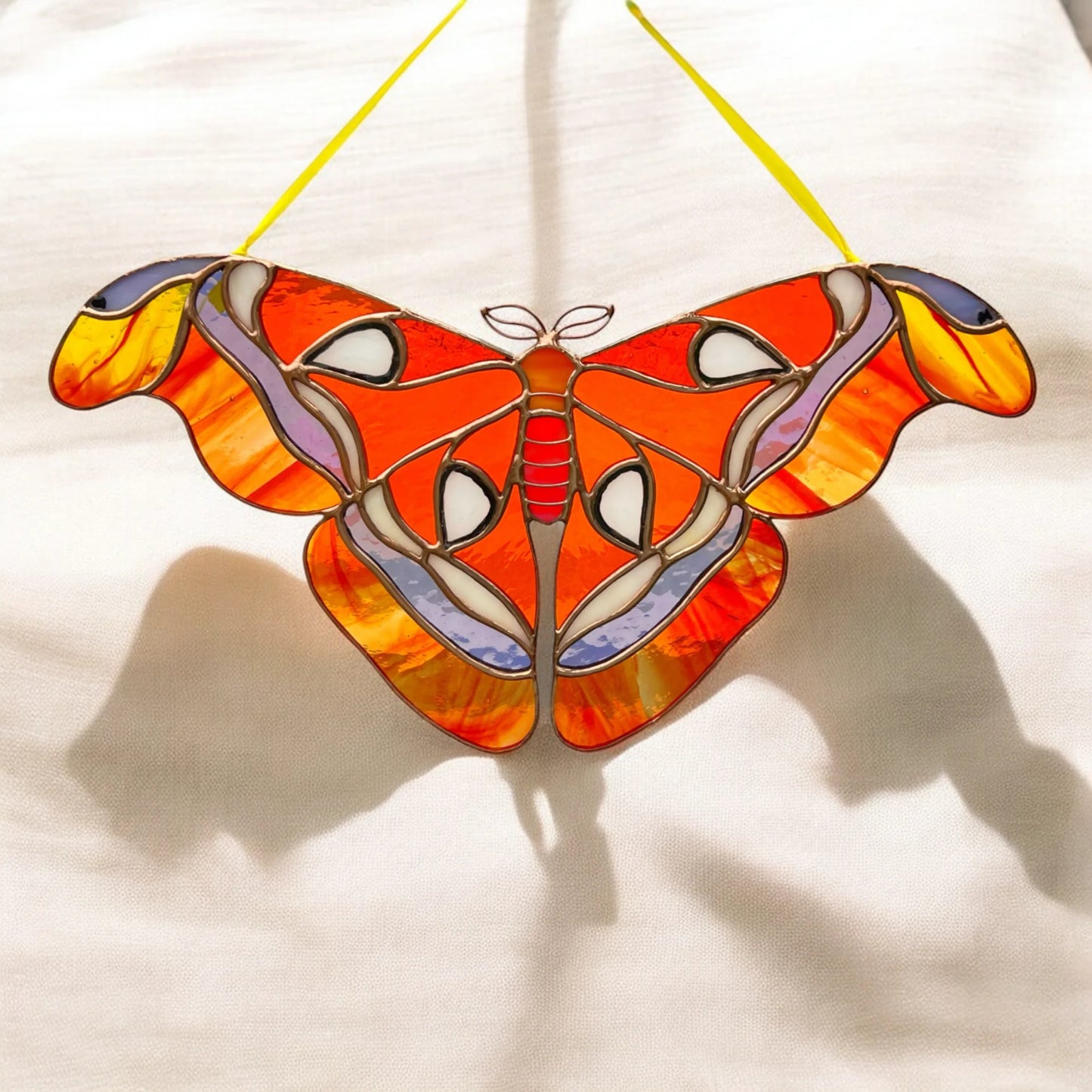 A stained glass moth suncatcher with vibrant orange, yellow, red, and purple sections, suspended by two yellow strings against a white fabric background with soft shadows.