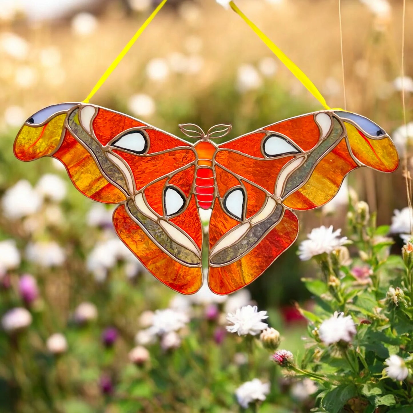A stained glass garden suncatcher shaped like a large moth, featuring vibrant orange, yellow, red, and white patterns with metallic silver outlines, suspended by a yellow string in a blurred outdoor garden setting.
