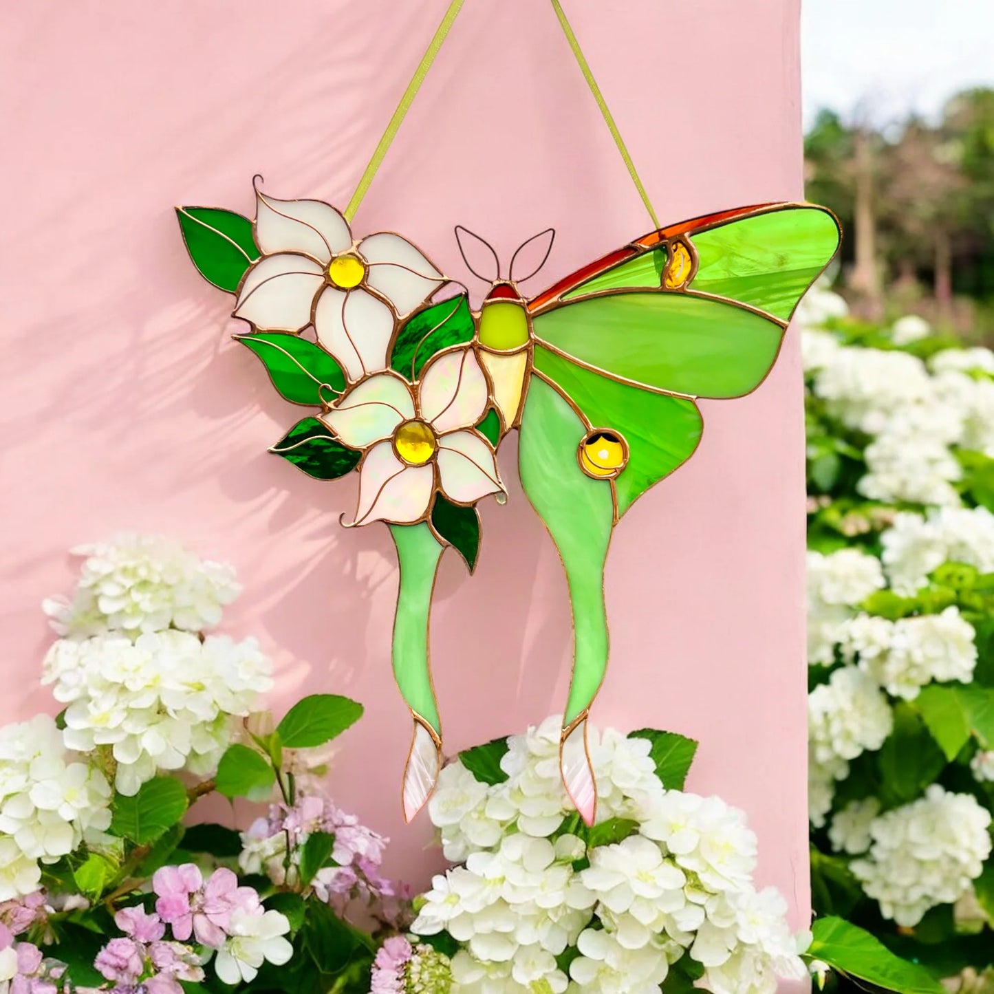 A stained glass butterfly suncatcher with green wings, white flowers, and green leaves, hanging against a pink wall with white flowers in the foreground.
