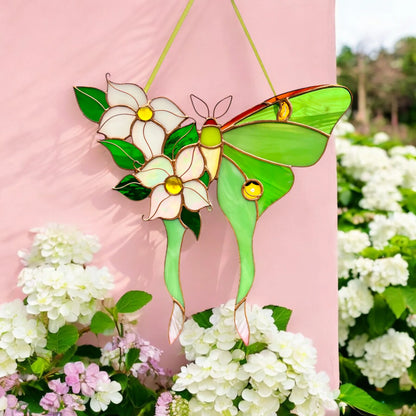 A stained glass butterfly suncatcher with green wings, white flowers, and green leaves, hanging against a pink wall with white flowers in the foreground.
