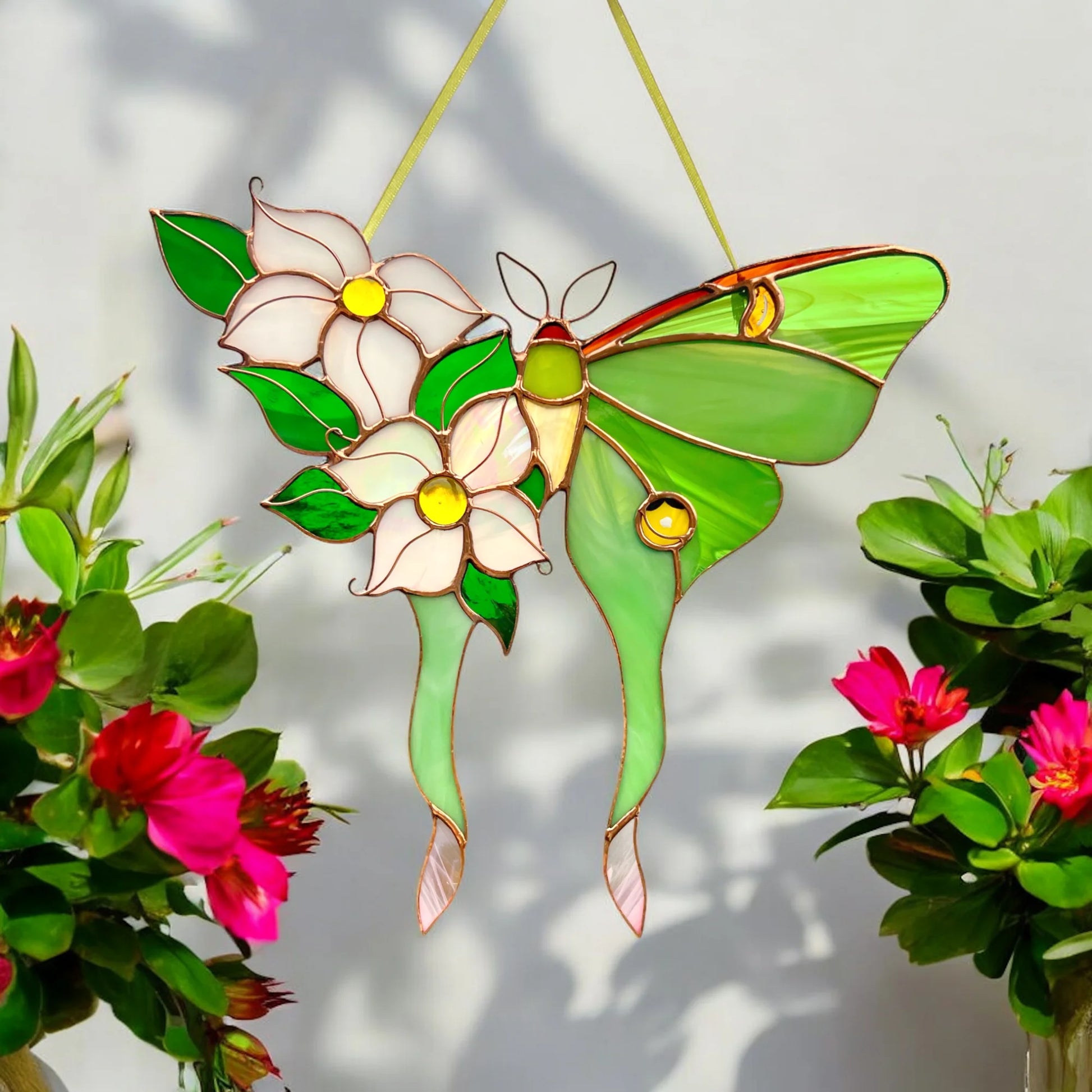 A stained glass hanging ornament shaped like a green moth with two white flowers and green leaves, suspended against a light wall with shadows, flanked by potted pink flowers.