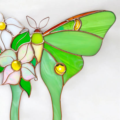 A stained glass suncatcher art piece featuring a green moth butterfly with yellow accents and white and pink flowers, set against a plain white background.