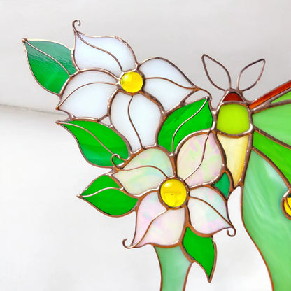 Close-up of a stained glass floral design featuring white and pale pink flowers with bright yellow centers, green leaves, and a green-and-yellow butterfly, outlined in copper-toned metal.