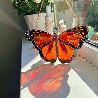 A stained-glass butterfly ornament with orange, red, black, and green sections hangs on a windowsill, casting a warm orange and red shadow on the white surface below.