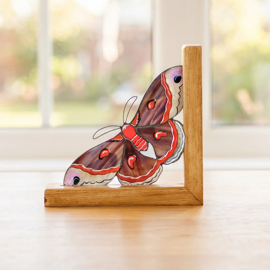 A gorgeous red stained glass cecropia moth bookend placed on a sunny windowsill, making a wonderful gift for book lovers.