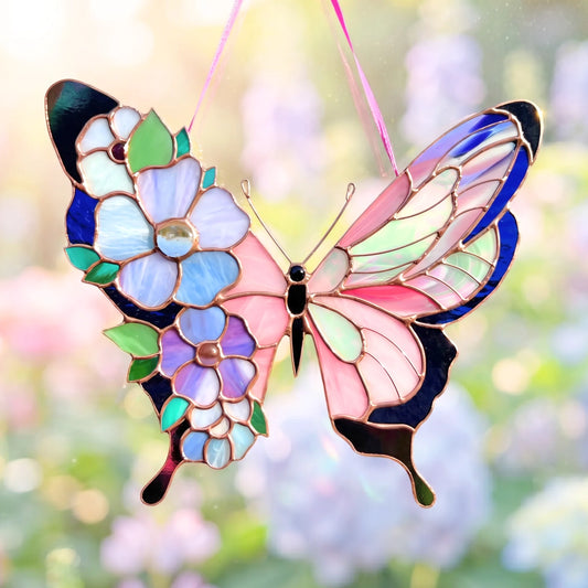 A stained glass butterfly suncatcher ornament with floral details, featuring pink, blue, purple, and green glass panels outlined in gold-toned metal, suspended by a pink ribbon against a soft, blurred garden background.