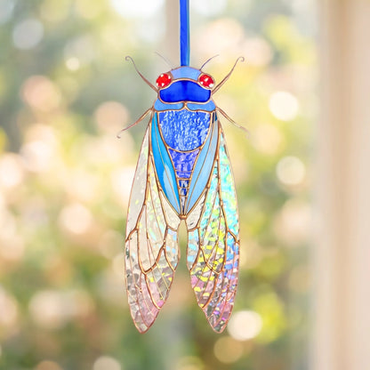 A stained glass cicada ornament hanging from a blue string, featuring iridescent wings, a blue body, and red eyes against a soft, blurred green and gold bokeh background.
