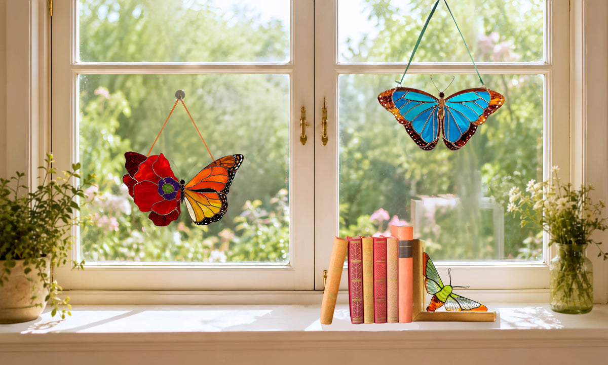 Two colorful stained glass butterfly window decorations hang on a white double-glass window with a garden view and one moth bookend standing pn a windowshell