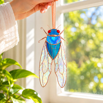 Hand hanging a blue stained glass cicada window hanging in a bright window beside a houseplant, light shining through its iridescent stained glass wings