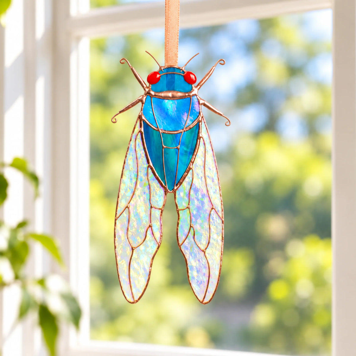 Blue stained glass cicada suncatcher with red glass eyes and iridescent stained glass cicada wings, hanging in a sunny window as handmade stained glass window decor
