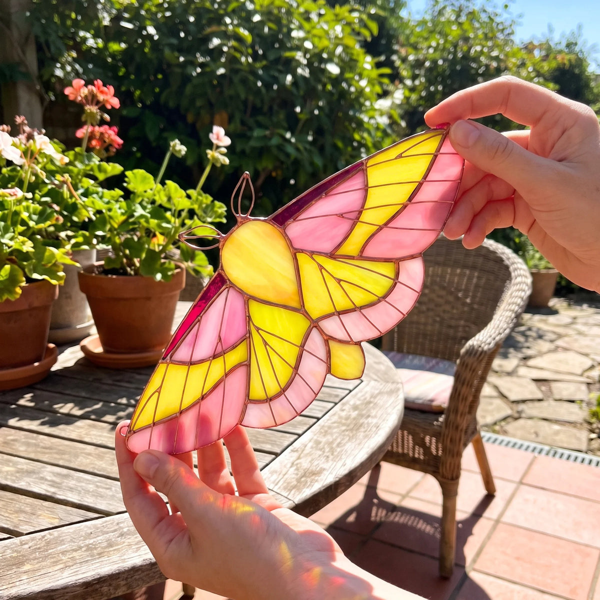 A stained-glass moth ornament with pink, yellow, and orange details is being held by two hands outdoors on a wooden table in a garden patio setting.