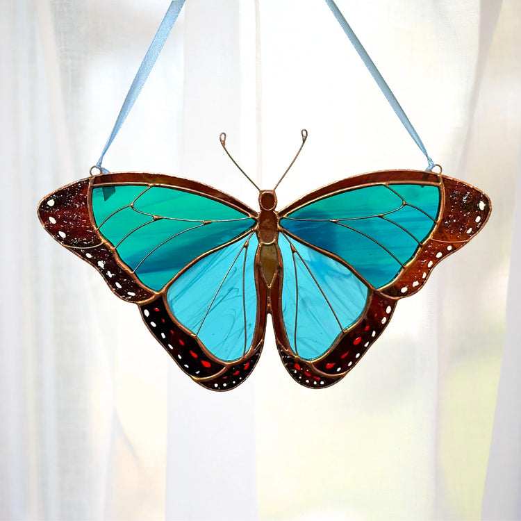 A morpho butterfly hanging ornament with bright turquoise and blue wings, brown borders, and small red spots, suspended by a light blue ribbon in front of a softly lit white curtain.