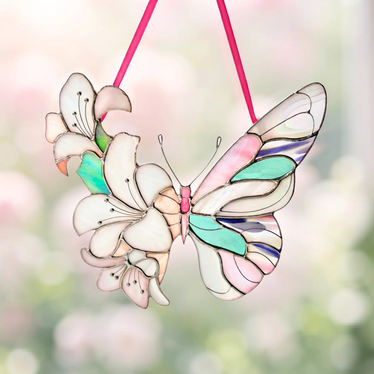 A stained glass butterfly ornament with white lily flowers, hanging from a pink ribbon against a soft, blurred green and pink background.