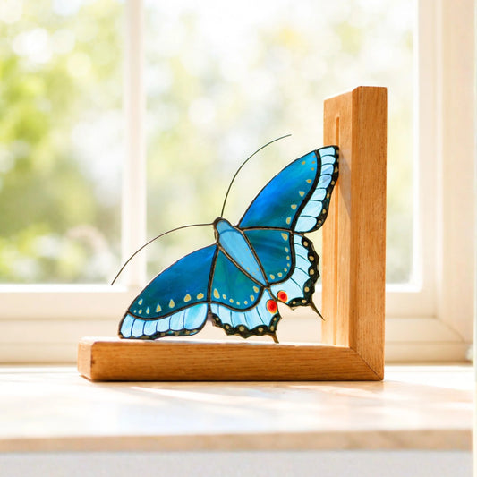 A stained glass butterfly bookend with bright blue wings, black outlines, and small red and yellow accents, mounted on a light wood L-shaped frame and placed on a windowsill with a blurred outdoor background.