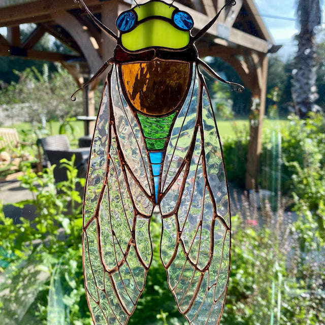 A stained glass cicada ornament with a bright yellow-green head, blue eyes, amber-brown body, and detailed green and blue wings, hanging in front of a window with a garden and wooden structure in the background.