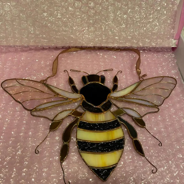 A stained glass bumblebee ornament with a black and yellow striped body, clear wings, and gold-toned metal framing, displayed on pink bubble wrap.