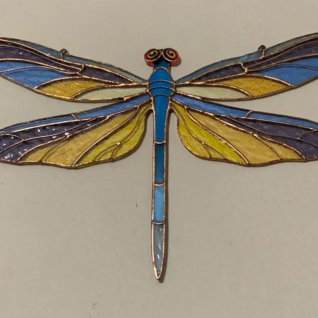 A stained glass dragonfly ornament with a bright blue body, yellow and blue wings, and gold-toned metal outlines, shown against a plain light background.