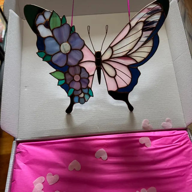 A stained-glass butterfly ornament with floral details in purple, blue, green, and pink, displayed inside an open white box with bright pink tissue paper and heart-shaped pieces scattered on top.