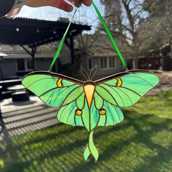 A stained glass luna moth ornament hanging in front of a window, with a hand holding the top string. The background shows a blurred backyard with grass, trees, and a house.