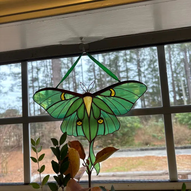 A stained glass luna moth ornament hanging in front of a window, with green wings, yellow accents, and dark outlines, set against an outdoor view of trees.