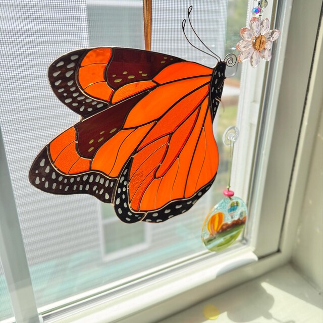 A stained glass monarch butterfly ornament hanging on a window, with orange and black wings, a small flower at the top, and a hanging glass charm below.