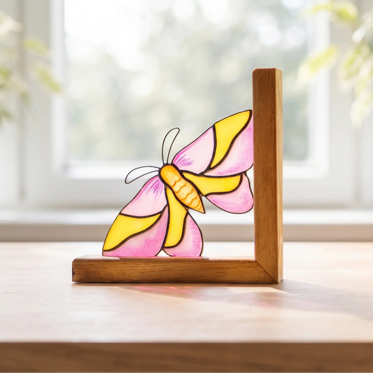 A stained-glass rosy maple moth ornament with pink and yellow wings, mounted on a wooden bokend on a tabletop in front of a softly blurred window.